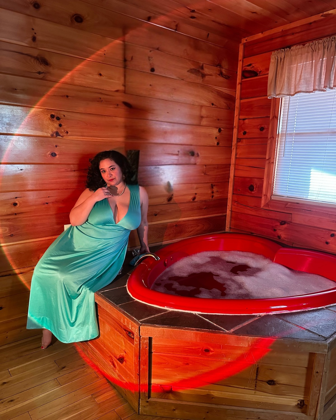 Romance author Corola De Rosa lounges beside a heart-shaped bathtub filled with water and bubbles. She wears a light blue vintage nightgown.